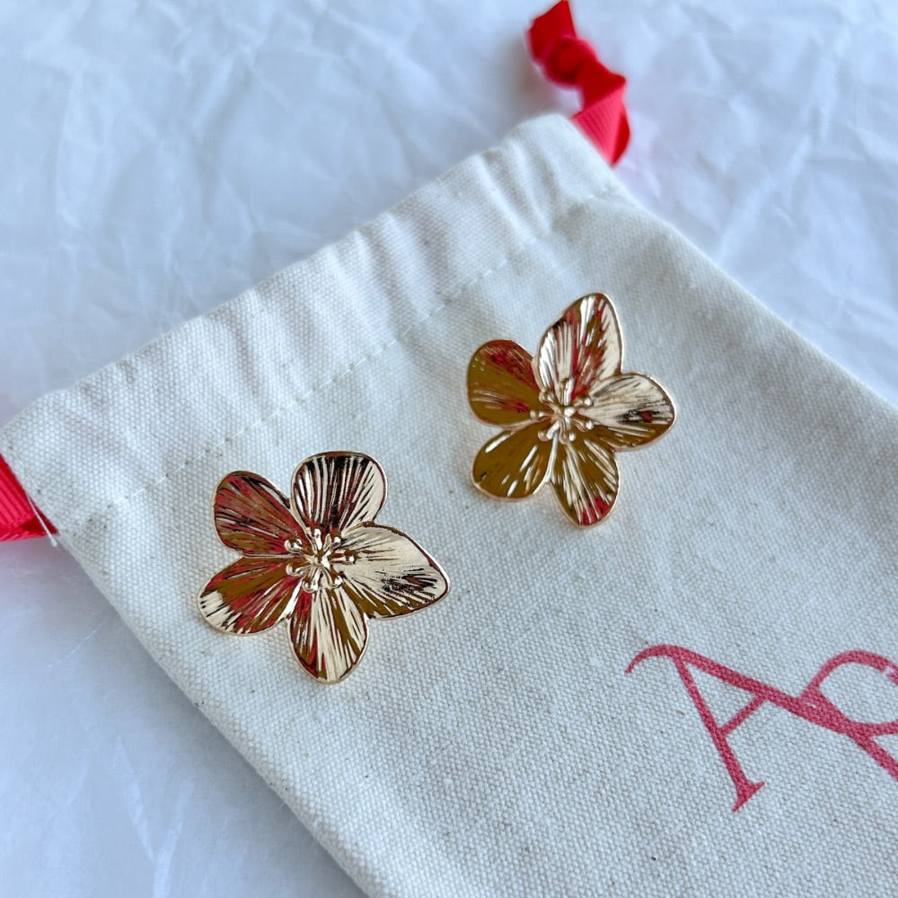 Aros Marsella en acabado dorado texturizado con detalles en relieve sobre bolsa de tela beige.