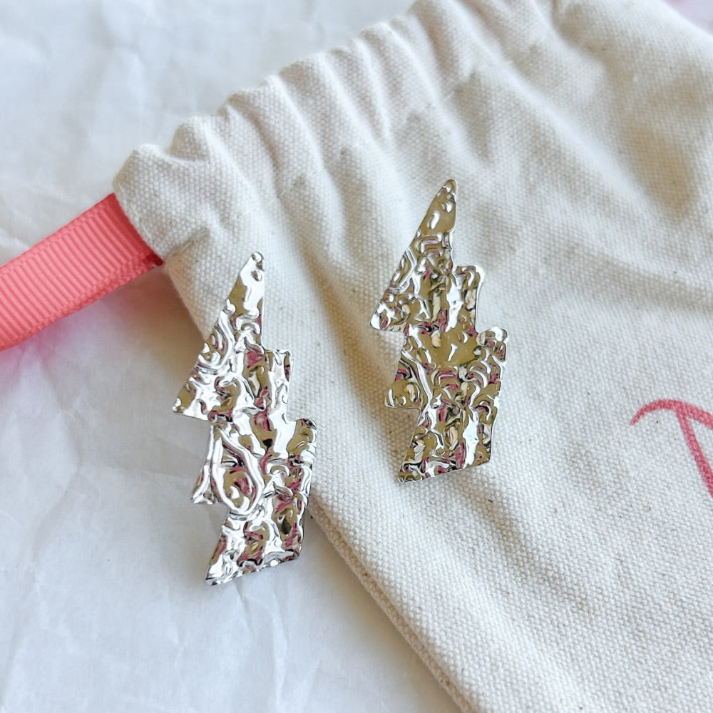 Aros de Plata Milán con diseño texturizado geométrico, exhibidos sobre una bolsa de tela clara con detalles en rojo.