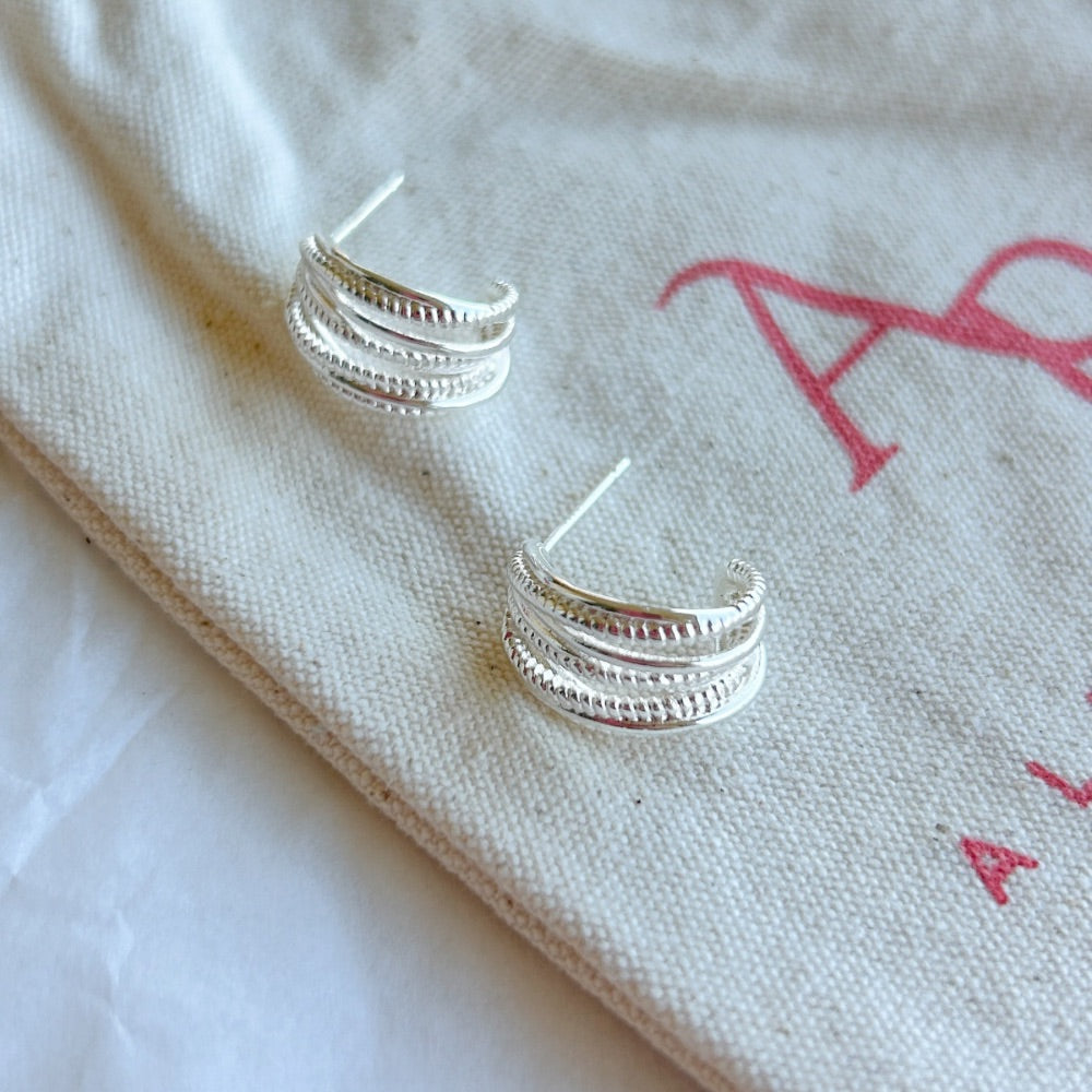 Aros de Plata Múnich con diseño de triple aro texturizado, exhibidos sobre una bolsa de tela clara con detalles en rojo.