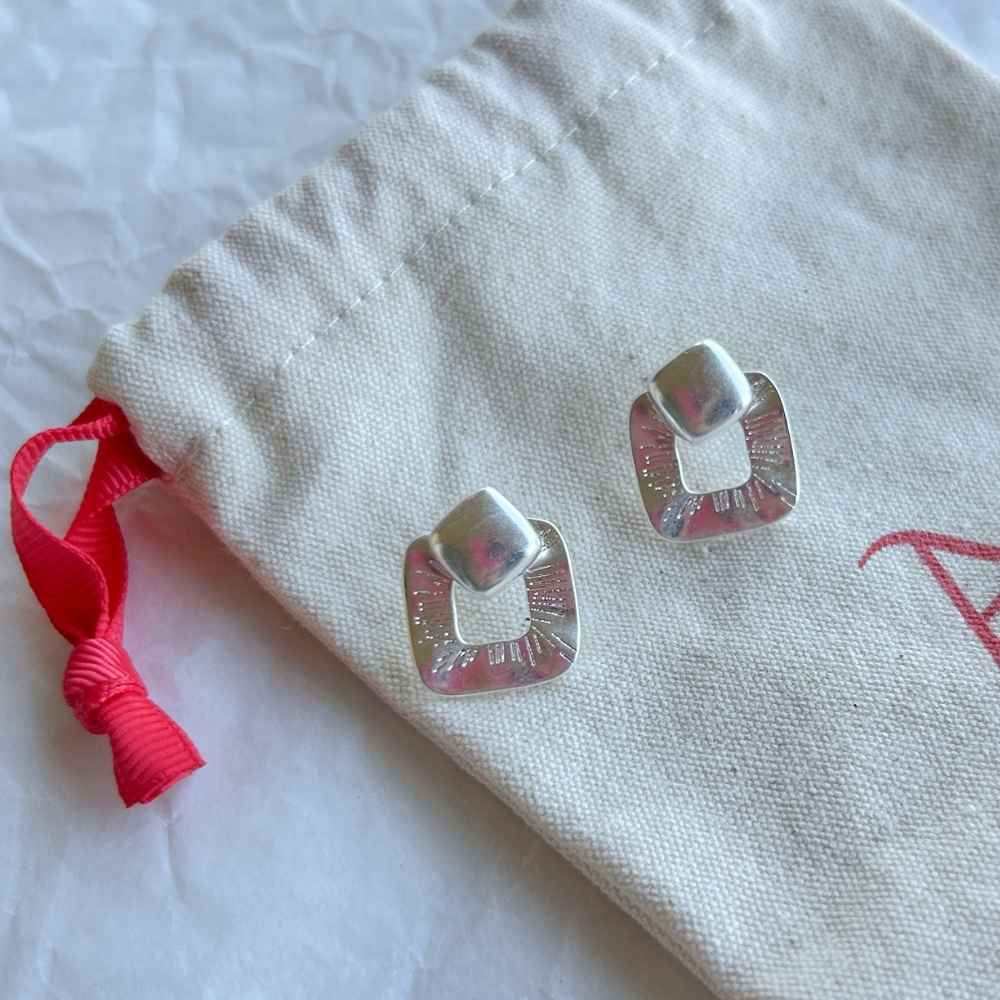 Aros de Plata Oslo con diseño cuadrado abierto y detalles grabados, exhibidos sobre una bolsa de tela clara con lazo rojo.