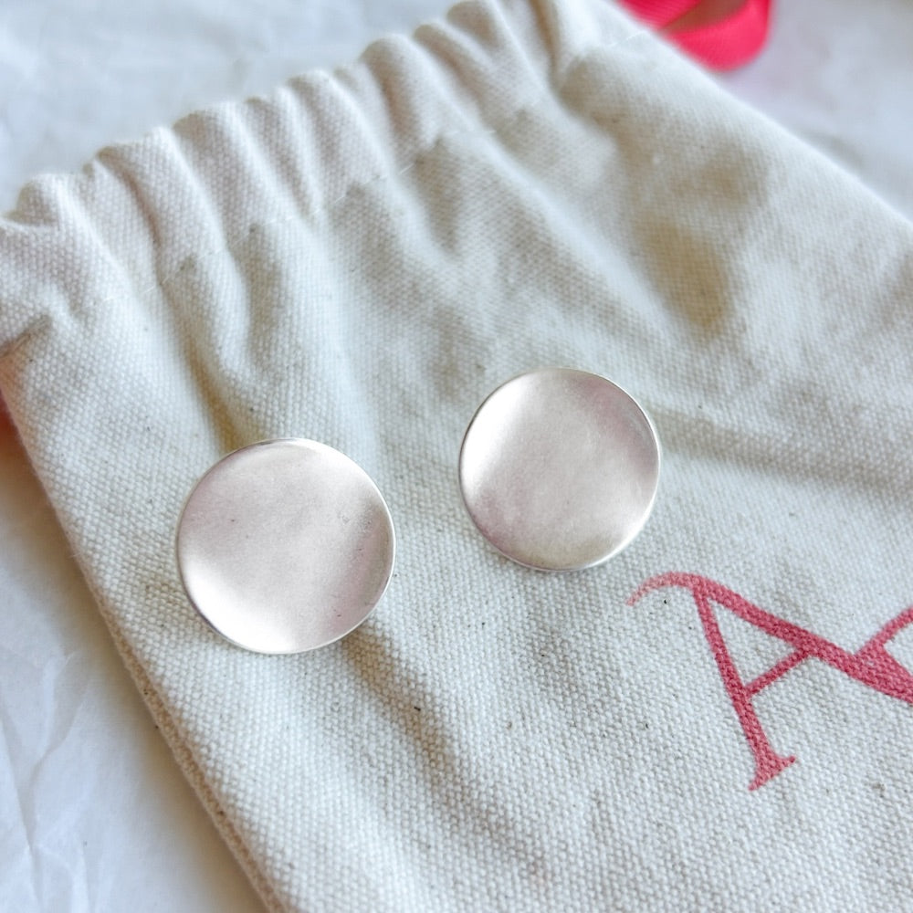 Aros de Plata Roma con diseño circular y acabado mate, exhibidos sobre una bolsa de tela clara con lazo rojo.