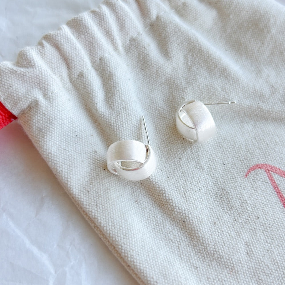 Aros de plata Zurich con diseño entrelazado y textura satinada sobre bolsa de tela.