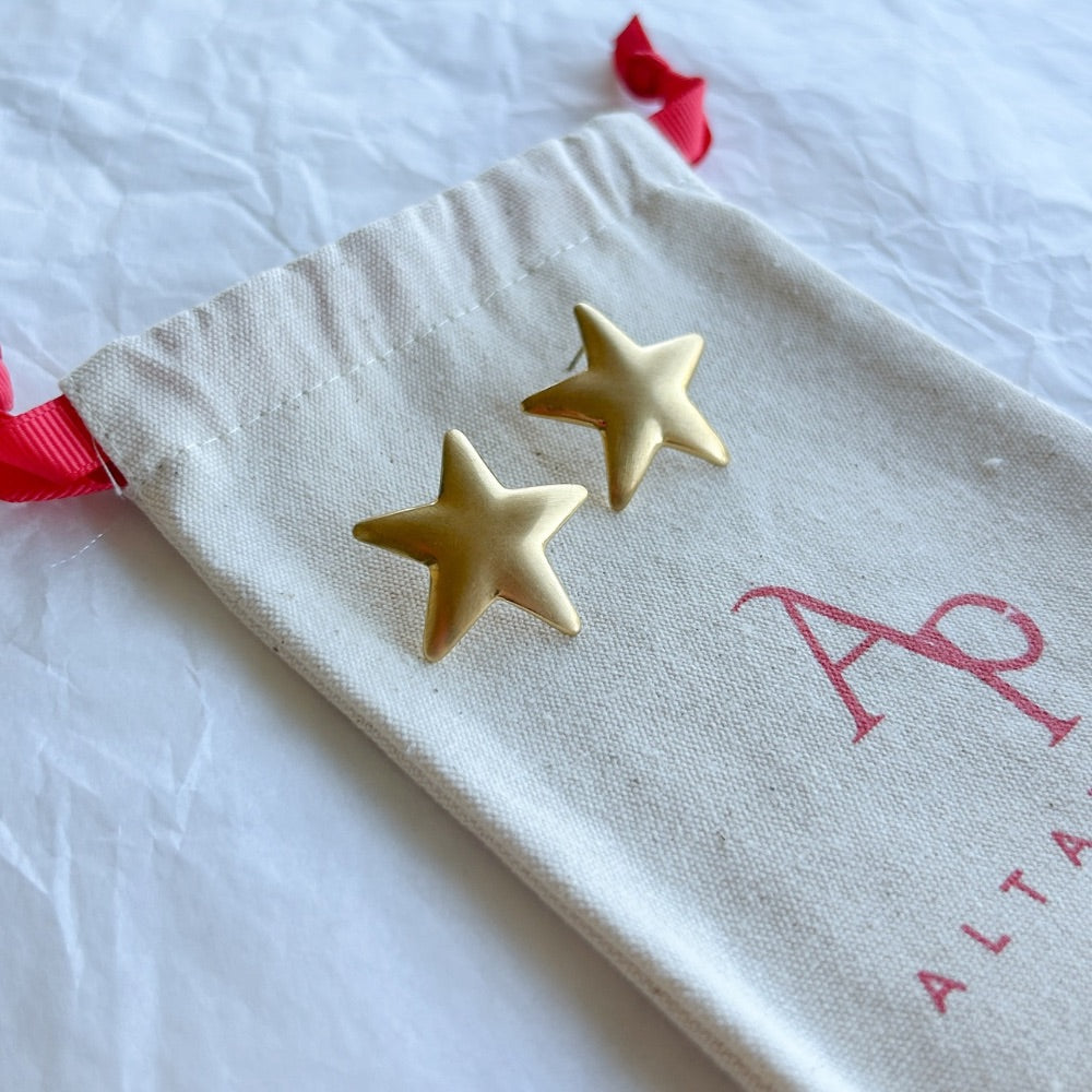 Aros Dorados Atenas con diseño de estrella y acabado mate, exhibidos sobre una bolsa de tela Altapinta.