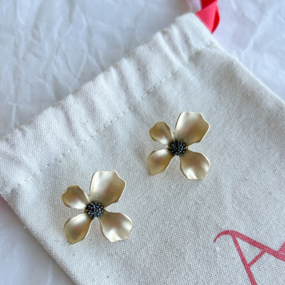 Aros París en baño de oro con pétalos satinados y centro texturizado sobre bolsa de tela.