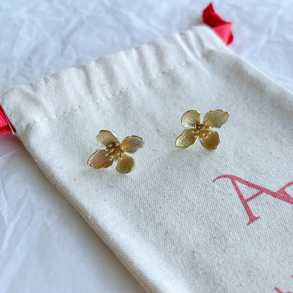Aros Dorados Patras con diseño de flor texturizada y acabado dorado, exhibidos sobre una bolsa de tela clara con lazo rojo.