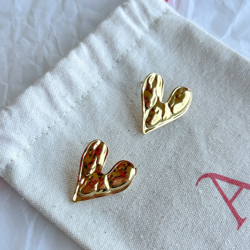 Aros Dorados Praga con diseño de corazón asimétrico y textura martillada, exhibidos sobre una bolsa de tela clara con lazo rojo.