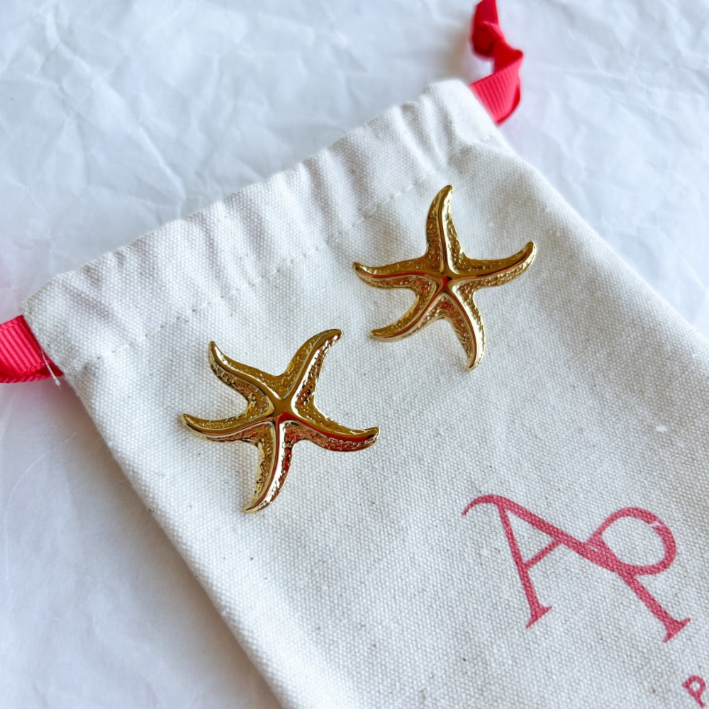 Aros Dorados Tampere con forma de estrella de mar y textura detallada, exhibidos sobre una bolsa de tela clara con lazo rojo.