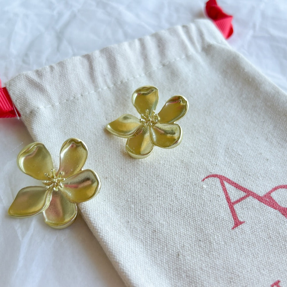 Aros Dorados Varsovia con diseño floral de pétalos curvados y centro en relieve, exhibidos sobre una bolsa de tela clara con lazo rojo.