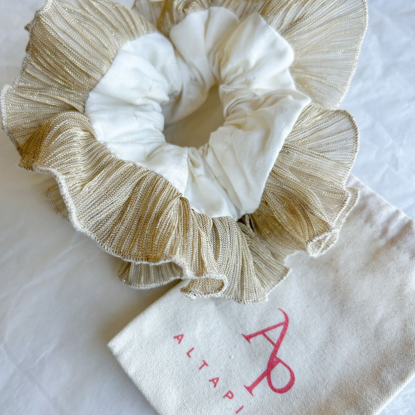 Bamba Doble Dorada Glow con plisado dorado y lazo blanco, presentada sobre una bolsa de tela decorativa.