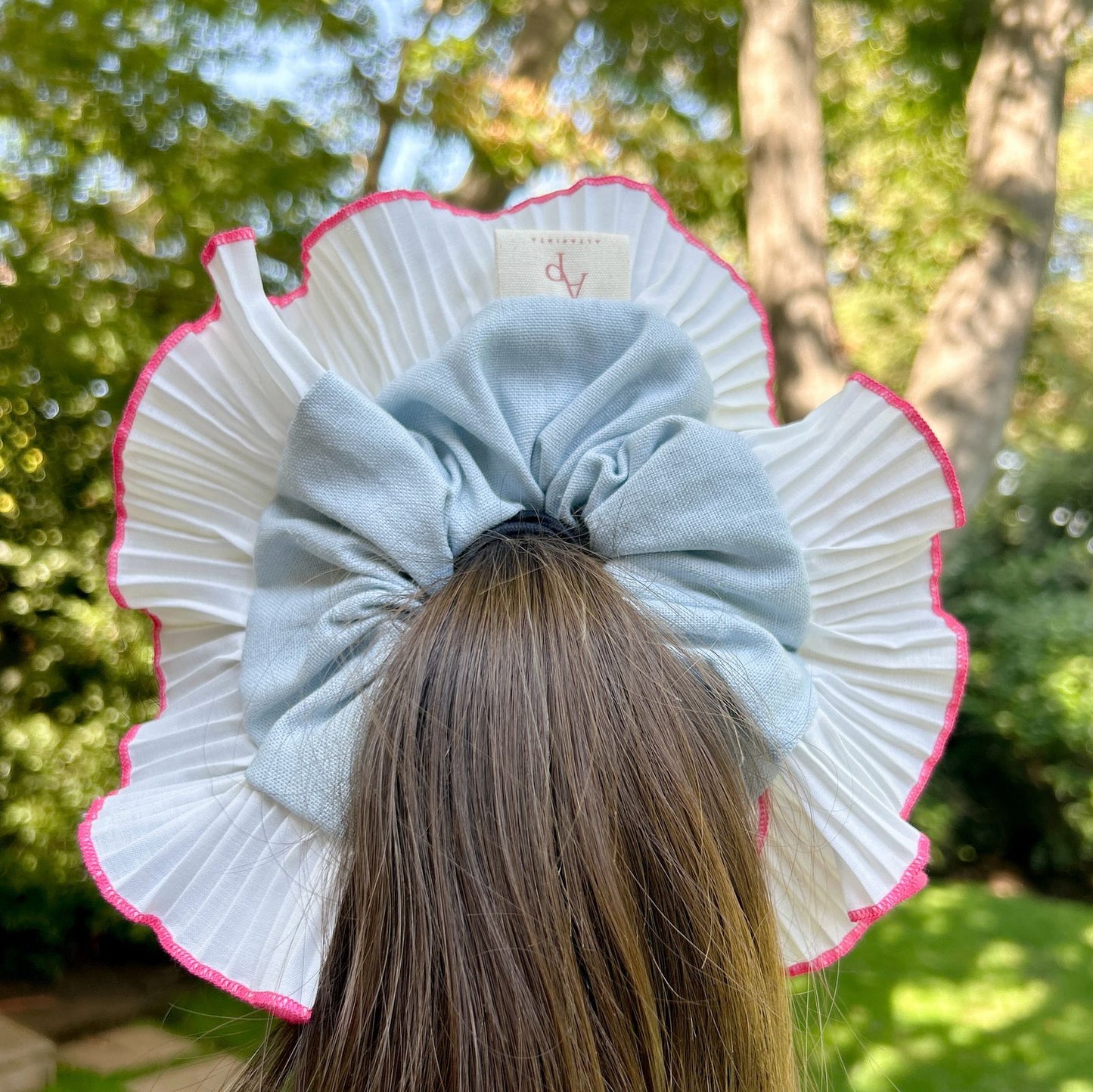 Bamba Simple Puerto con lazo azul claro y plisado blanco con borde rosa, decorando una coleta alta en un entorno natural.