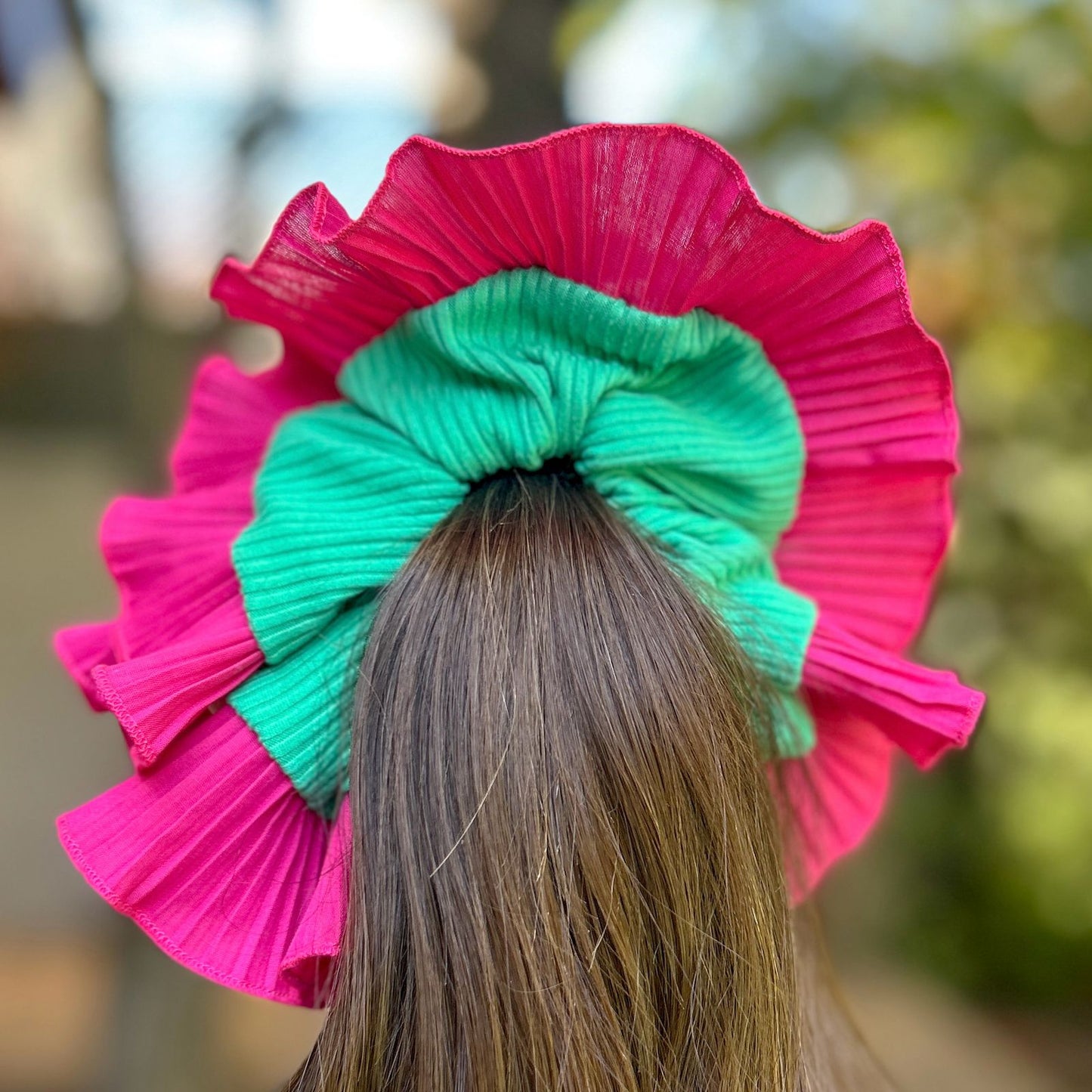 Bamba Simple Tropical con lazo verde intenso y plisado fucsia, decorando una coleta alta con un fondo natural.