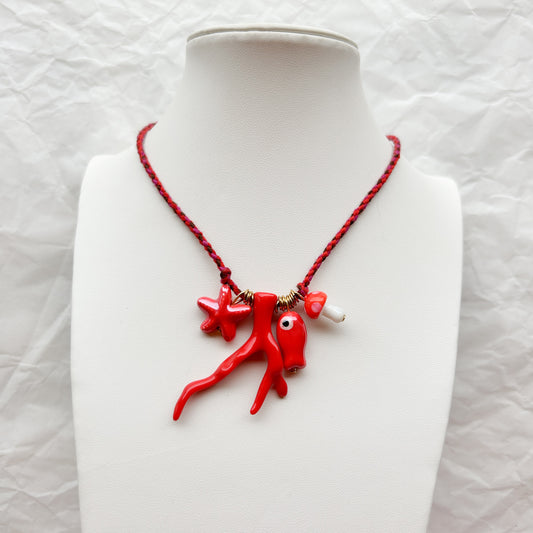 Collar Artesanal Coral trenzado en rojo y negro, con dijes de estrella de mar, ramas de coral y amuleto de ojo protector rojo, exhibido en un busto blanco.