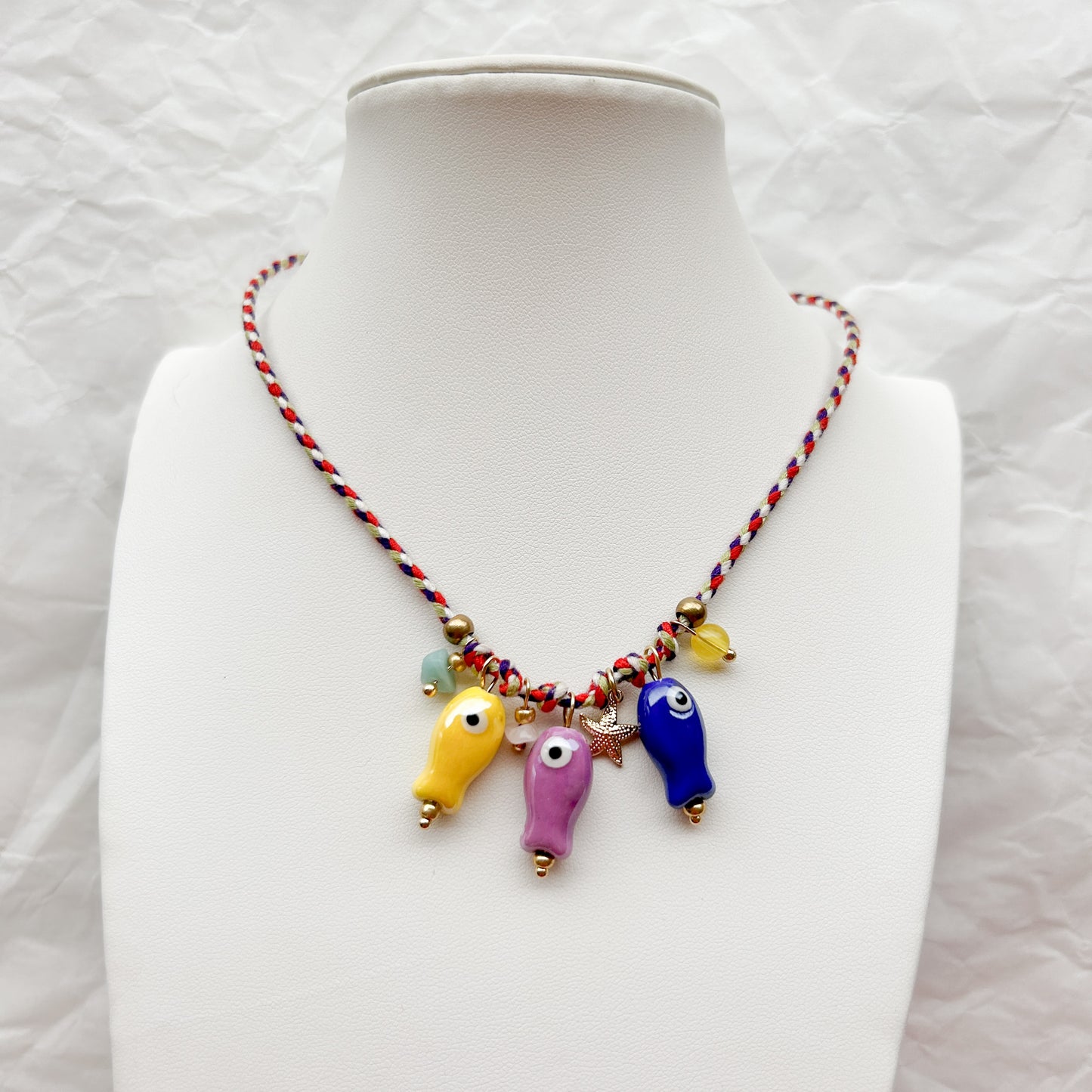 Collar Artesanal Olas con tejido trenzado en rojo, blanco y azul, decorado con dijes de peces de cerámica y detalles dorados, exhibido sobre un busto blanco.