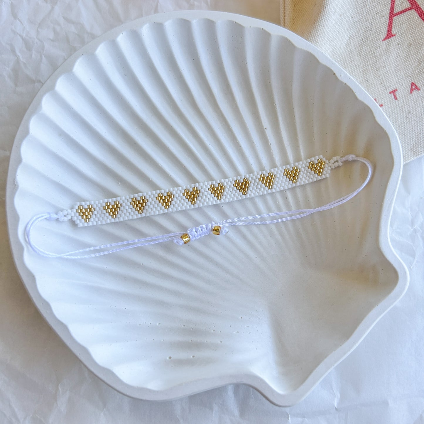 Pulsera Artesanal Corazones Catalina tejida a mano con mostacillas doradas sobre una base blanca, con cierre ajustable, presentada sobre una concha blanca.