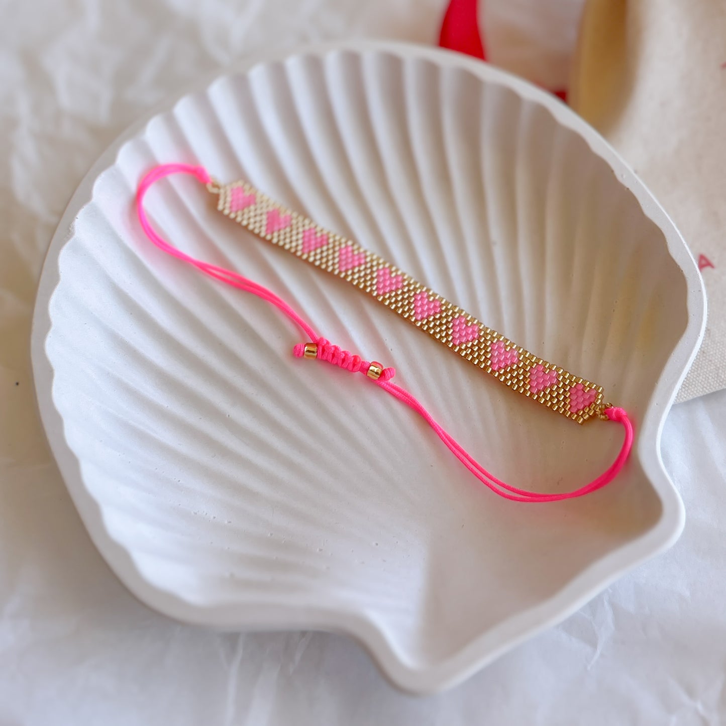 Pulsera Artesanal Corazones Ignacia en rosa y dorado sobre concha blanca, con diseño de corazones tejidos a mano.