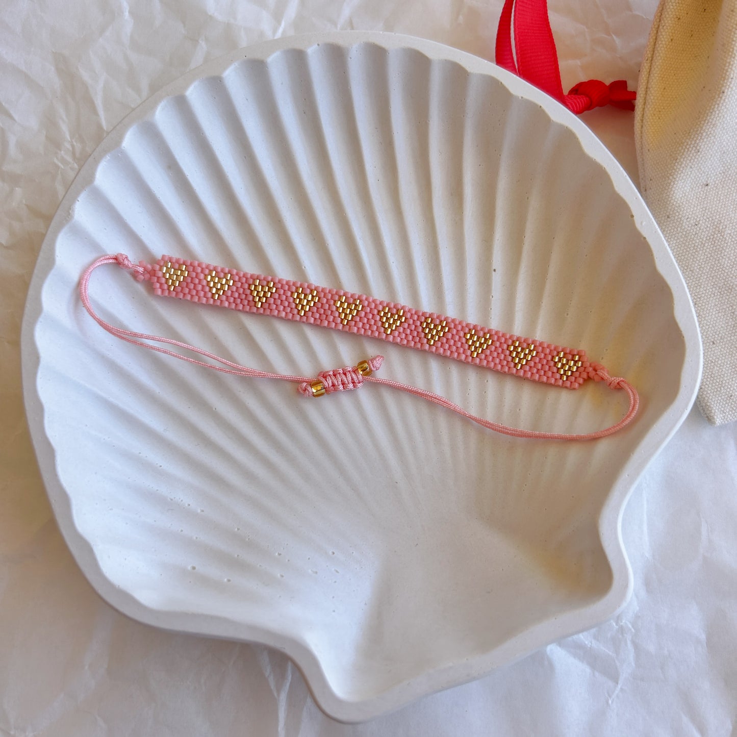 Pulsera artesanal tejida a mano con mostacillas rosa y corazones dorados, sobre una concha blanca.