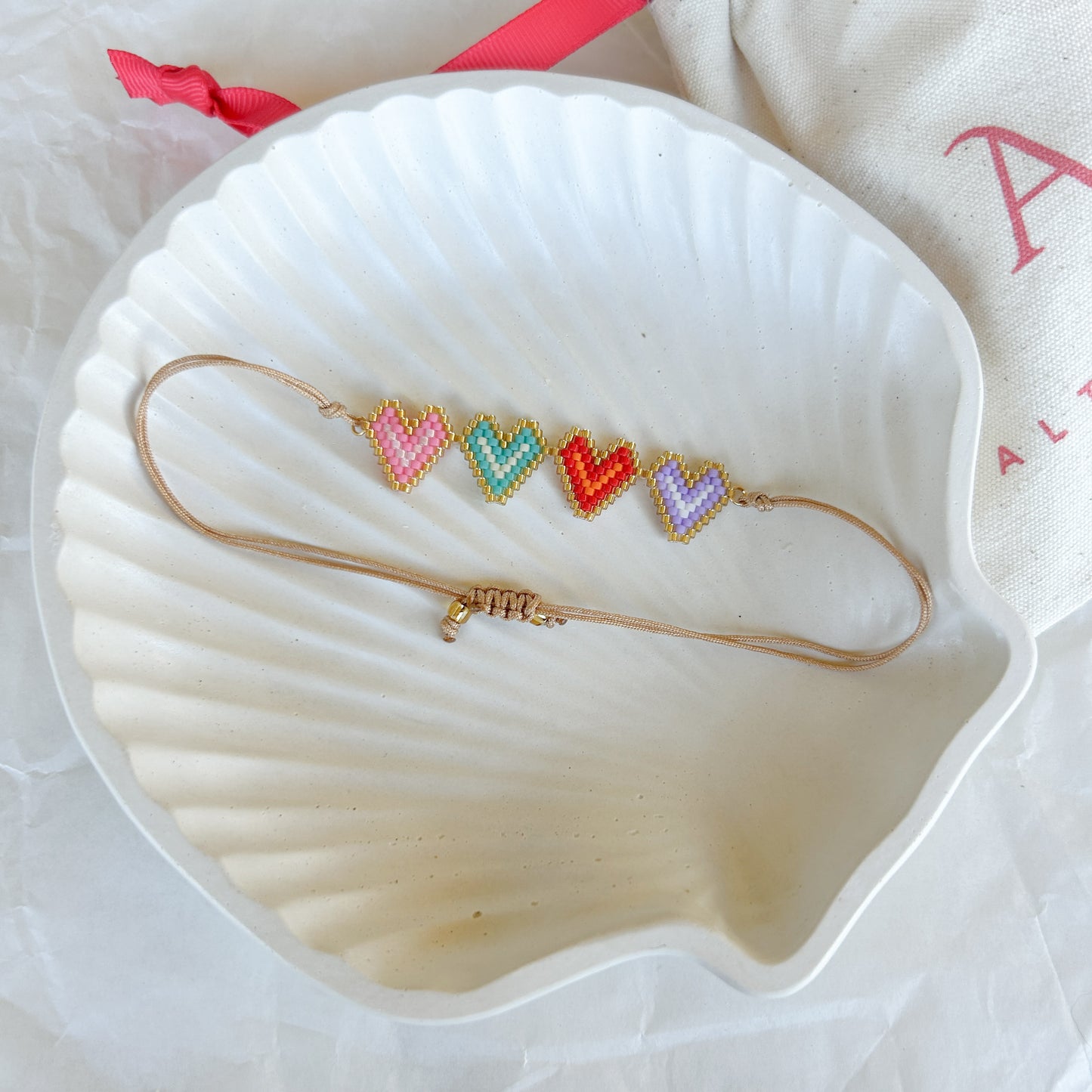 Pulsera artesanal con cuatro corazones tejidos en diferentes colores, enmarcados en dorado, sobre una concha blanca.