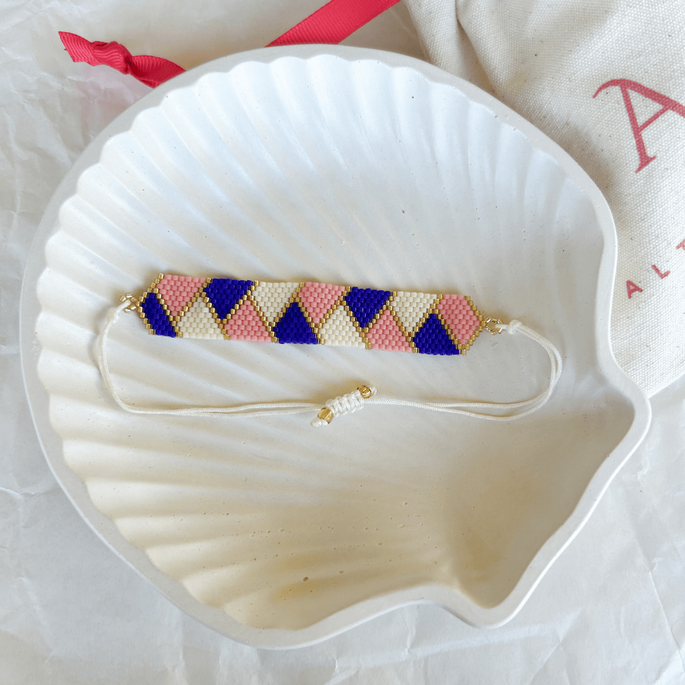 Pulsera Artesanal Tejida de Mostacillas Amanda sobre una concha blanca, mostrando su diseño de triángulos en azul, rosa y crema con detalles dorados y cierre ajustable.
