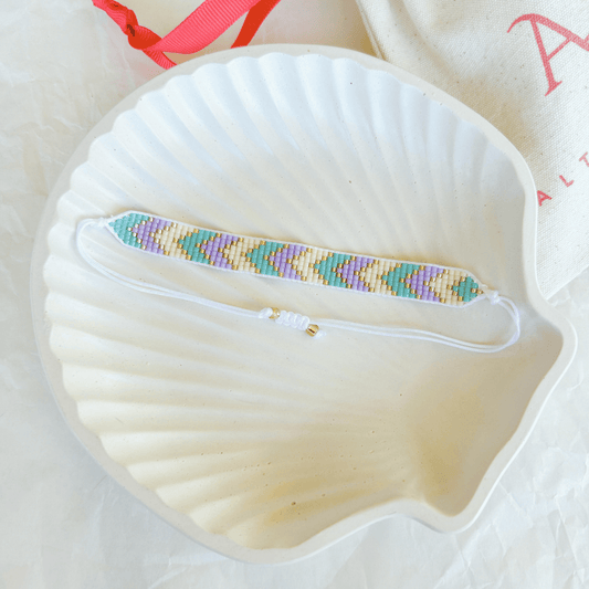 Pulsera Artesanal Tejida de Mostacillas Ángeles en blanco con detalles en lila y dorado, con un patrón geométrico.