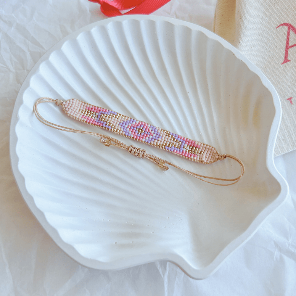 Pulsera Artesanal Tejida de Mostacillas Bernardita en tonos rosados y dorados, colocada sobre una concha blanca, resaltando su diseño detallado y cierre ajustable.