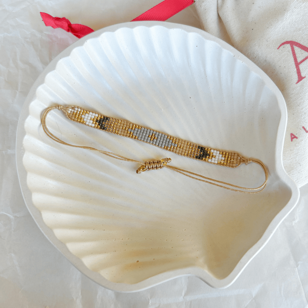 Pulsera Artesanal Tejida de Mostacillas Carolina en tonos dorados, negros y blancos, exhibida sobre una concha blanca, destacando su diseño geométrico y cierre ajustable.