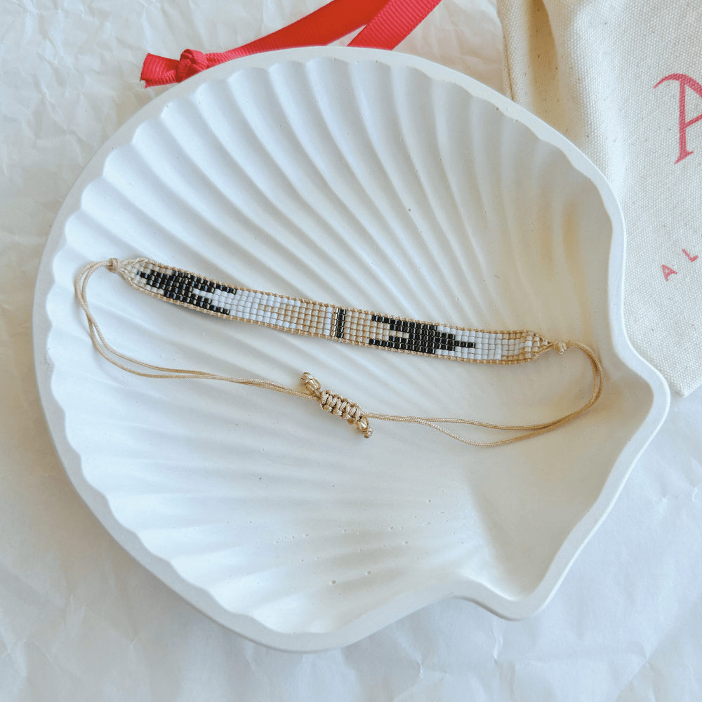 Pulsera Artesanal Tejida de Mostacillas Cristina en tonos blanco y oro, con detalles geométricos y cierre ajustable, sobre una concha blanca.