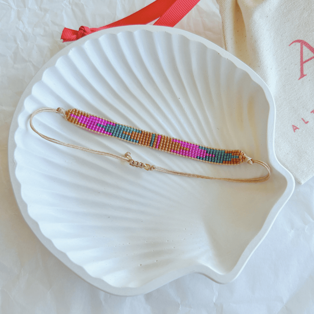 Pulsera Artesanal Tejida de Mostacillas Cristina en tonos rosa fucsia, naranja y dorado, con cierre ajustable, sobre una concha blanca.