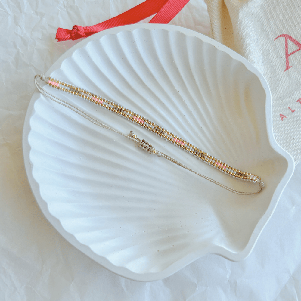 Pulsera Artesanal Tejida de Mostacillas Dominga en tonos cálidos y dorados con cierre ajustable, sobre una concha blanca.