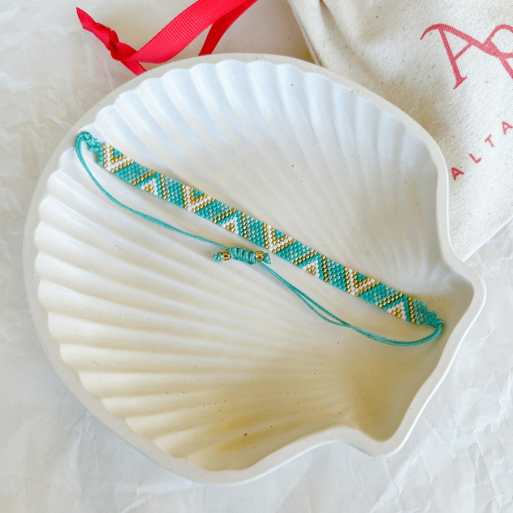 Pulsera Artesanal Tejida de Mostacillas Eloísa con diseño geométrico en tonos turquesa, dorado y blanco, con cierre ajustable, sobre una concha blanca.
