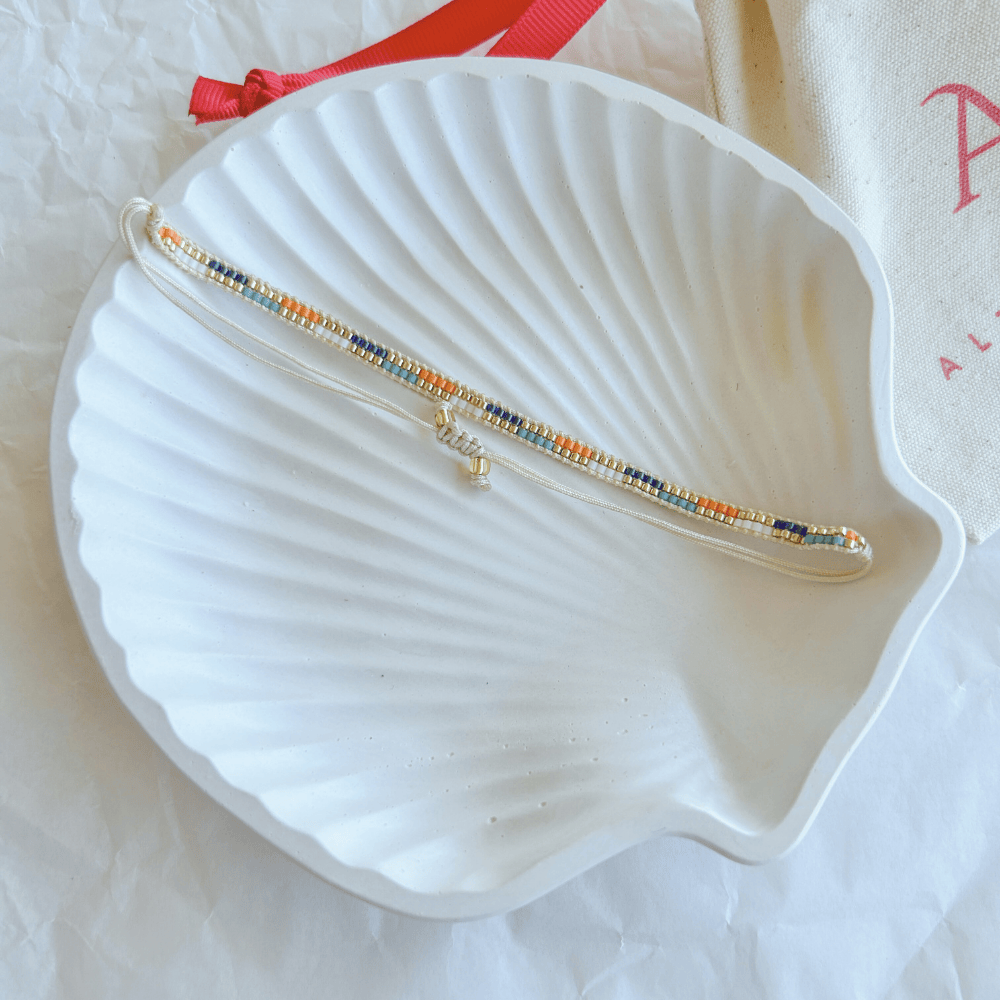 Pulsera Artesanal Tejida de Mostacillas Filippa en tonos azul, naranja y beige, con cierre ajustable, sobre una concha blanca. Diseño artesanal con mostacillas de alta calidad.