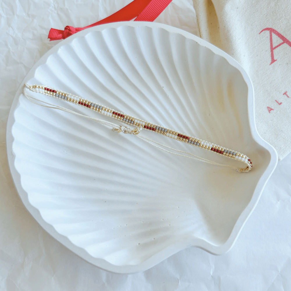 Pulsera Artesanal Tejida de Mostacillas Filippa en tonos rojo, dorado y beige, con cierre ajustable, sobre una concha blanca. Accesorio delicado y versátil.