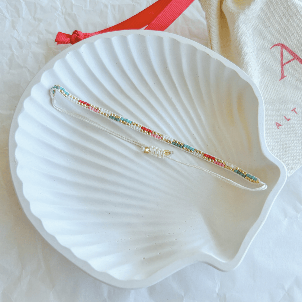 Pulsera Artesanal Tejida de Mostacillas Filippa en tonos turquesa, rojo y blanco, con cierre ajustable, sobre una concha blanca. Diseño hecho a mano con detalles dorados.