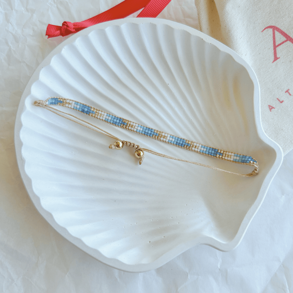 Pulsera Artesanal Tejida de Mostacillas Francisca en tonos azul y dorado, con cierre ajustable en hilo beige, sobre una concha blanca. Diseño hecho a mano con mostacillas de alta calidad.