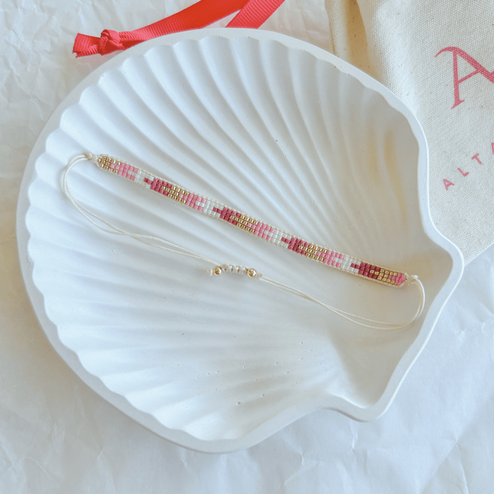 Pulsera Artesanal Tejida de Mostacillas Francisca en tonos rosa y dorado, con cierre ajustable en hilo blanco, sobre una concha blanca. Accesorio delicado y versátil con detalles artesanales.