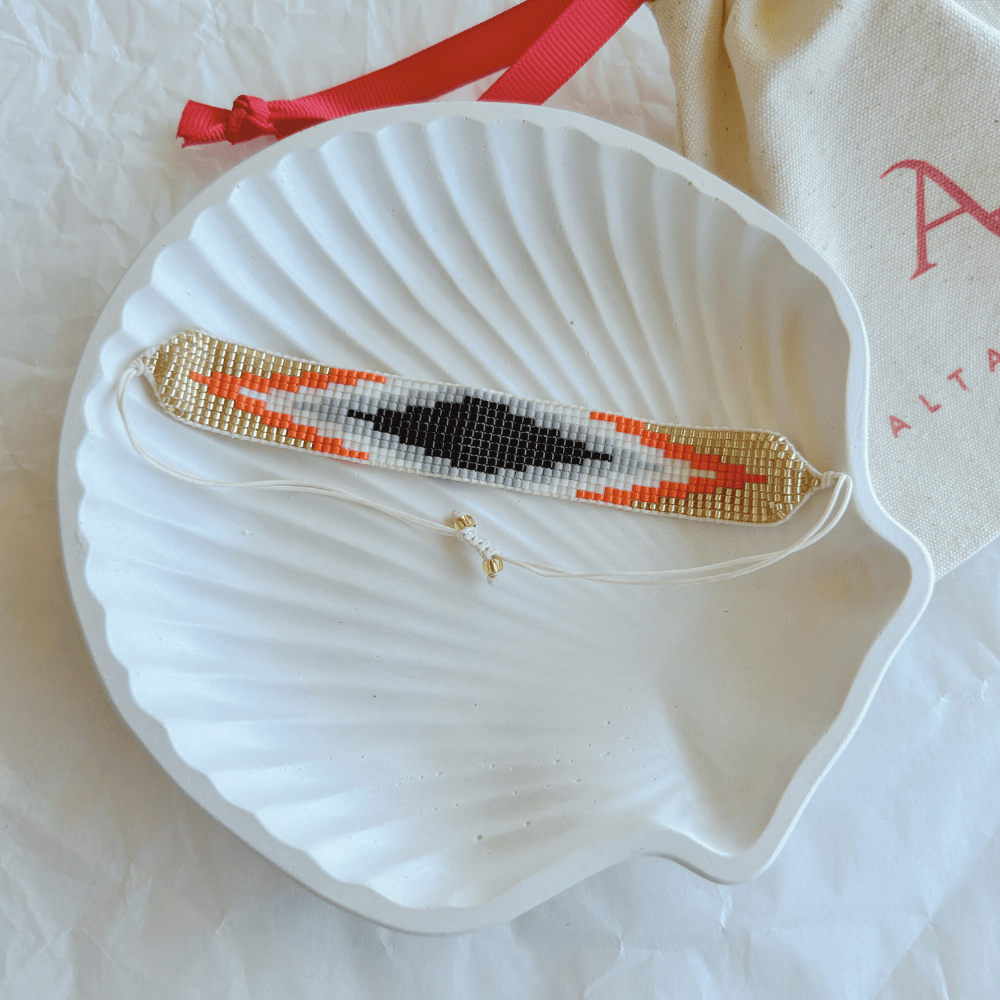Pulsera Artesanal Tejida de Mostacillas Loreto en tonos negro, blanco, naranja y dorado, con cierre ajustable en hilo blanco, sobre una concha blanca. Accesorio elegante y sofisticado.