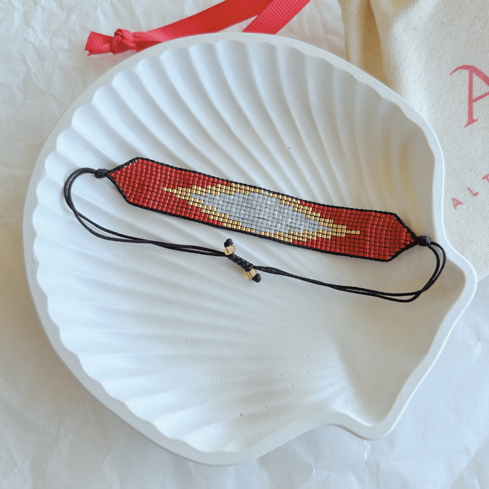 Pulsera Artesanal Tejida de Mostacillas Loreto en tonos rojo, gris y dorado, con cierre ajustable en hilo negro, sobre una concha blanca. Diseño geométrico artesanal.