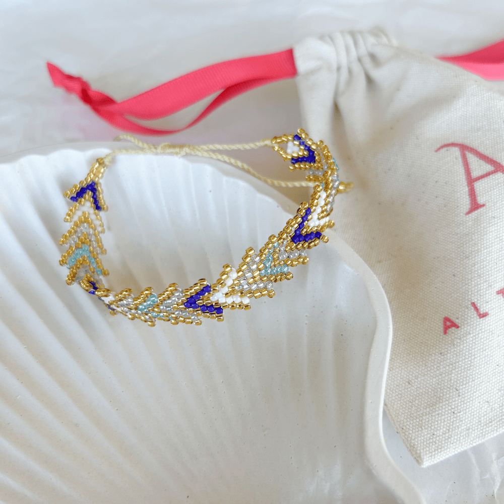 Pulsera Artesanal Tejida de Mostacillas María Gracia en tonos azul y blanco, con detalles dorados y diseño sofisticado, sobre una concha blanca.