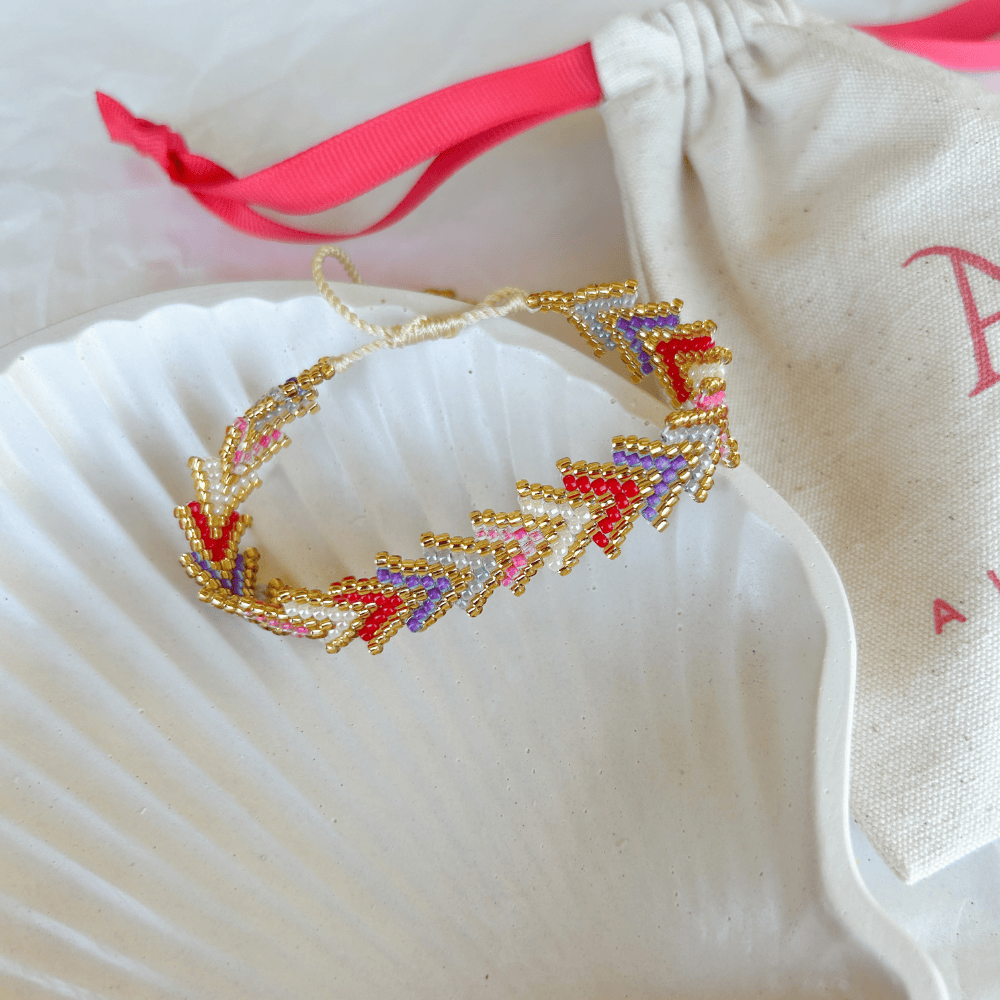 Pulsera Artesanal Tejida de Mostacillas María Gracia en tonos rojo y lila, con detalles dorados y diseño entrelazado, sobre una concha blanca.