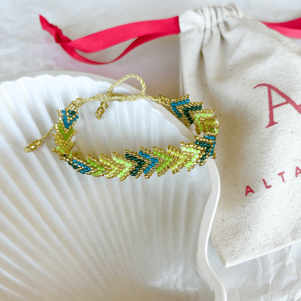 Pulsera Artesanal Tejida de Mostacillas María Gracia en tonos verde y azul, con detalles dorados y diseño trenzado, sobre una concha blanca.
