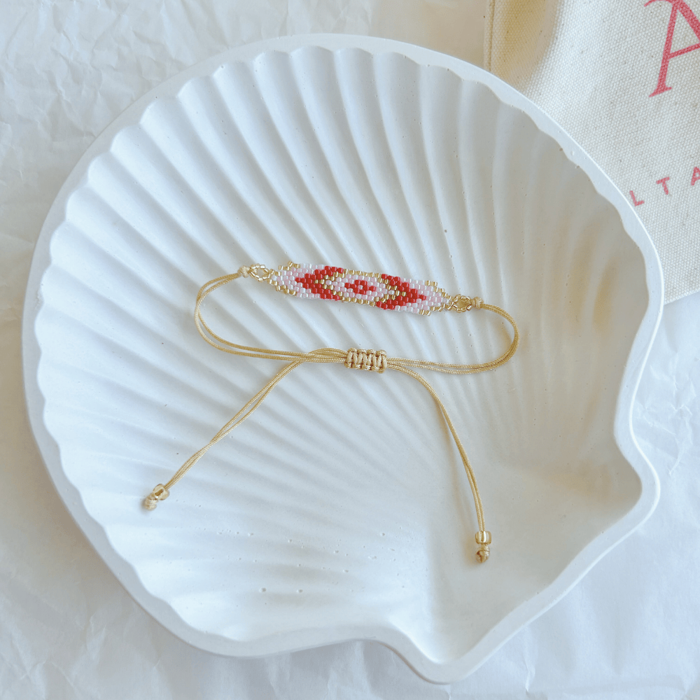 Pulsera Artesanal Tejida de Mostacillas Pola en tonos rojo y rosa, con diseño de ojo y cierre ajustable en hilo beige, sobre una concha blanca.