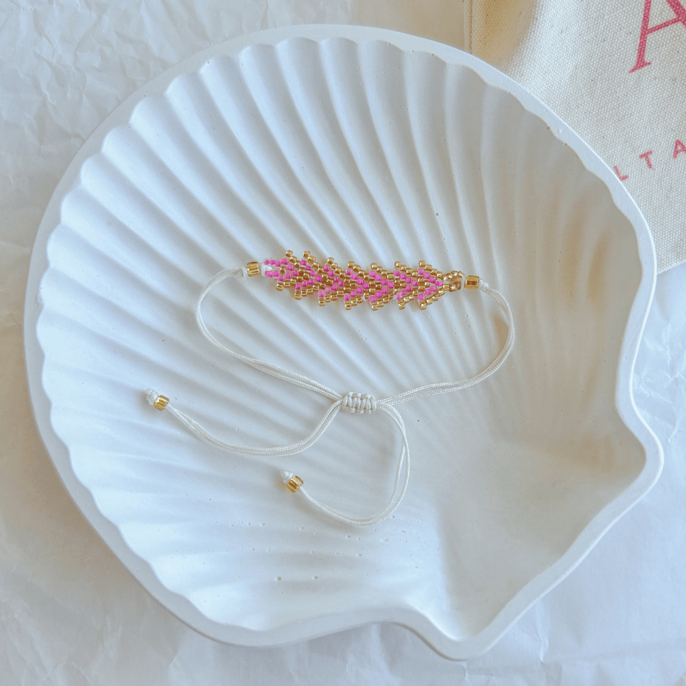 Pulsera Artesanal Tejida de Mostacillas Tere en rosa con dorado, con cierre ajustable en hilo beige, sobre una concha blanca.