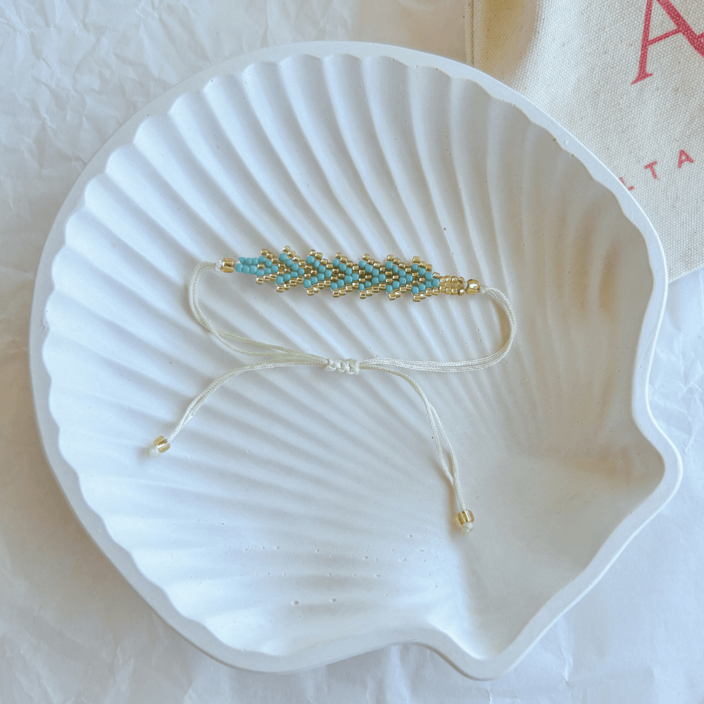Pulsera Artesanal Tejida de Mostacillas Tere en verde profundo con dorado, con cierre ajustable en hilo beige, sobre una concha blanca.