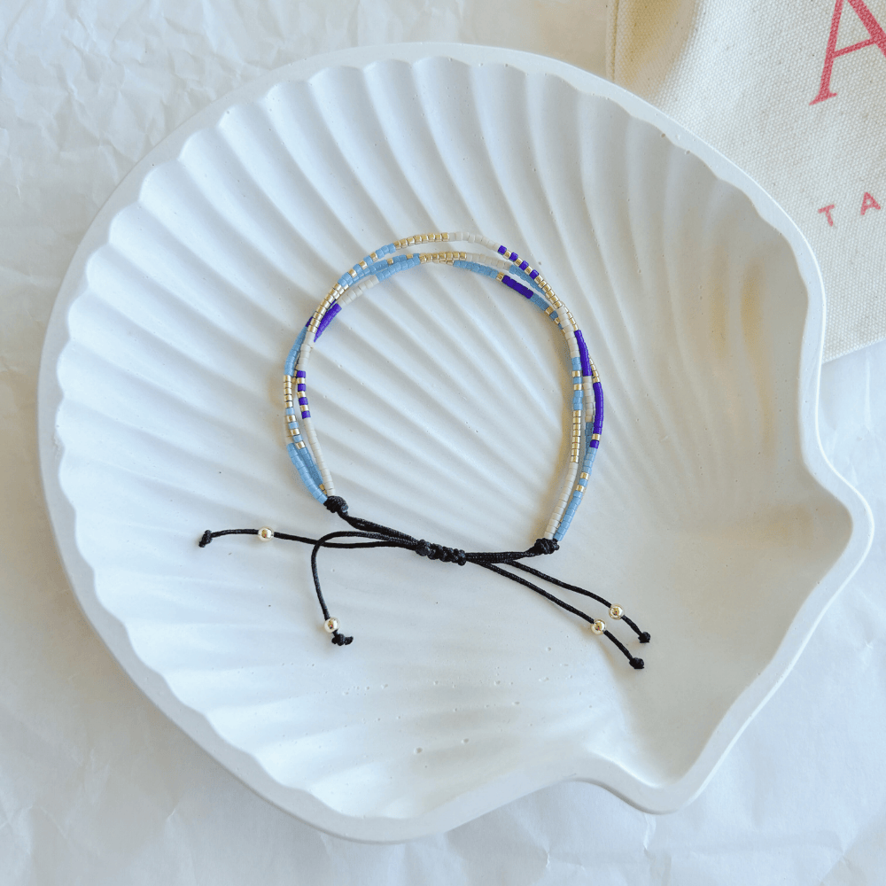 Pulsera Artesanal Triple Martina sobre una concha blanca, destacando su diseño de tres hileras en tonos azul, dorado y blanco con cierre ajustable.