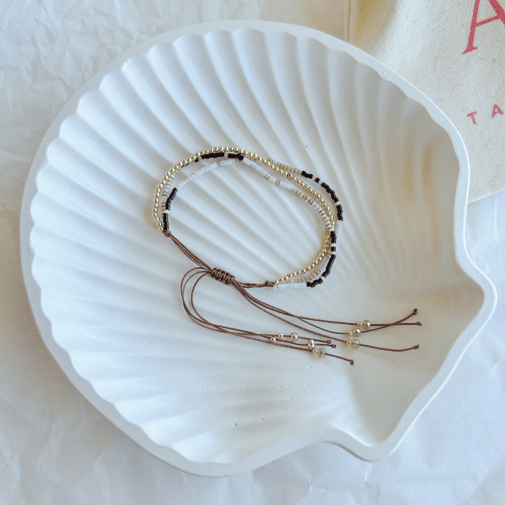 Pulsera Artesanal Triple Matilde sobre una concha blanca, resaltando su diseño de tres hileras en negro, dorado y blanco con cierre ajustable.