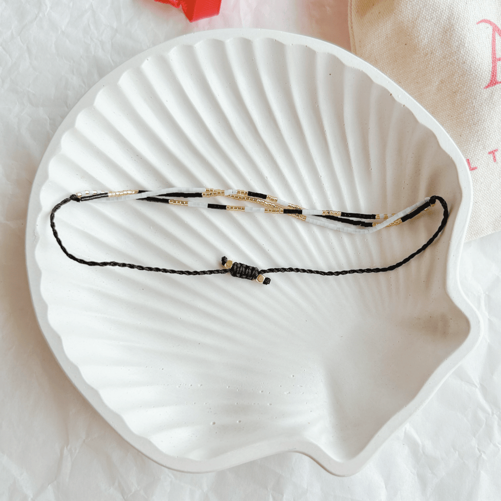 Pulsera Artesanal Triple Olimpia sobre una concha blanca, destacando su diseño de tres hileras en tonos blanco, negro y dorado con cierre ajustable.