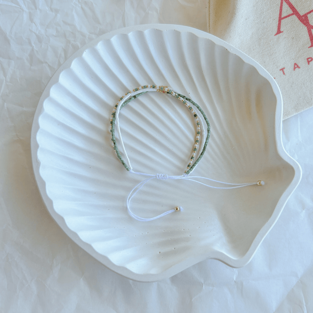 Pulsera Artesanal Triple Rafaela sobre una concha blanca, con un diseño artesanal de tres hileras en verde, blanco y dorado con cierre ajustable.