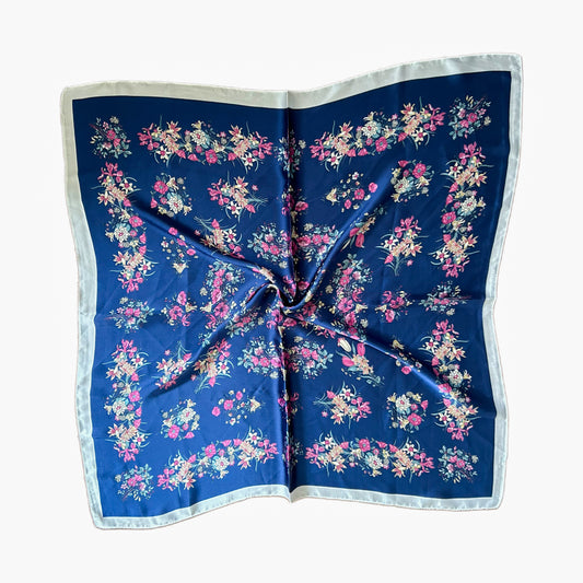 Pañuelo Calla con diseño floral en tonos claros sobre un fondo azul profundo, confeccionado en símil seda y viscosa, tamaño 70x70 cm. Elegante y versátil.