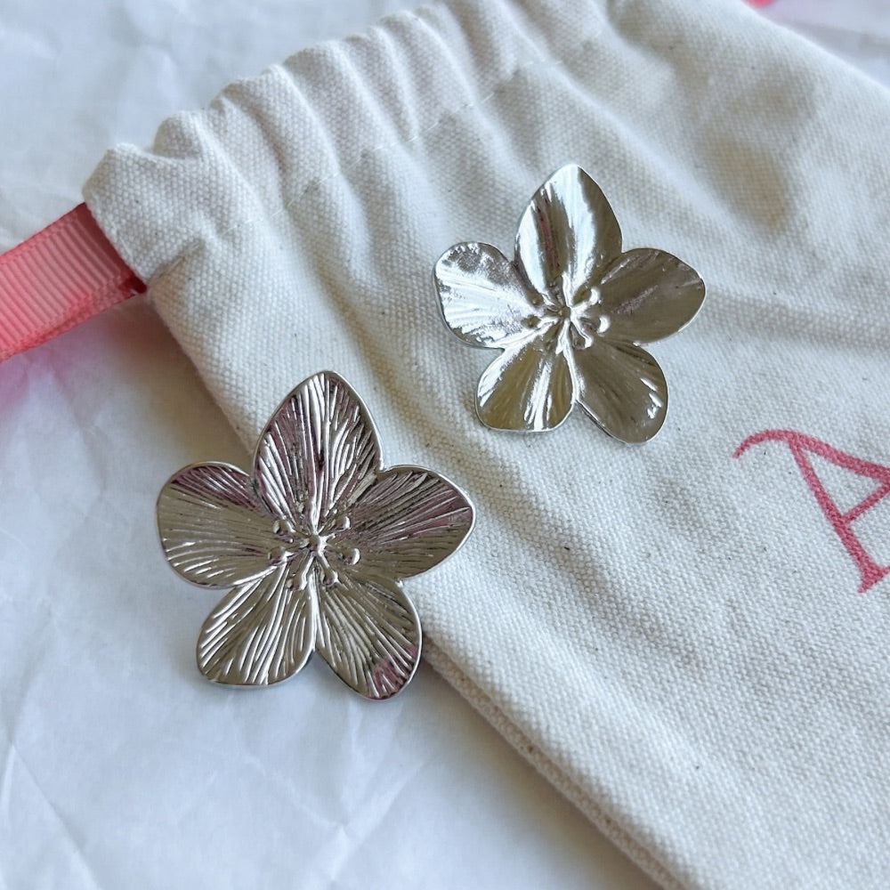 Aros Marsella en acabado plateado con diseño floral texturizado sobre bolsa de tela beige.