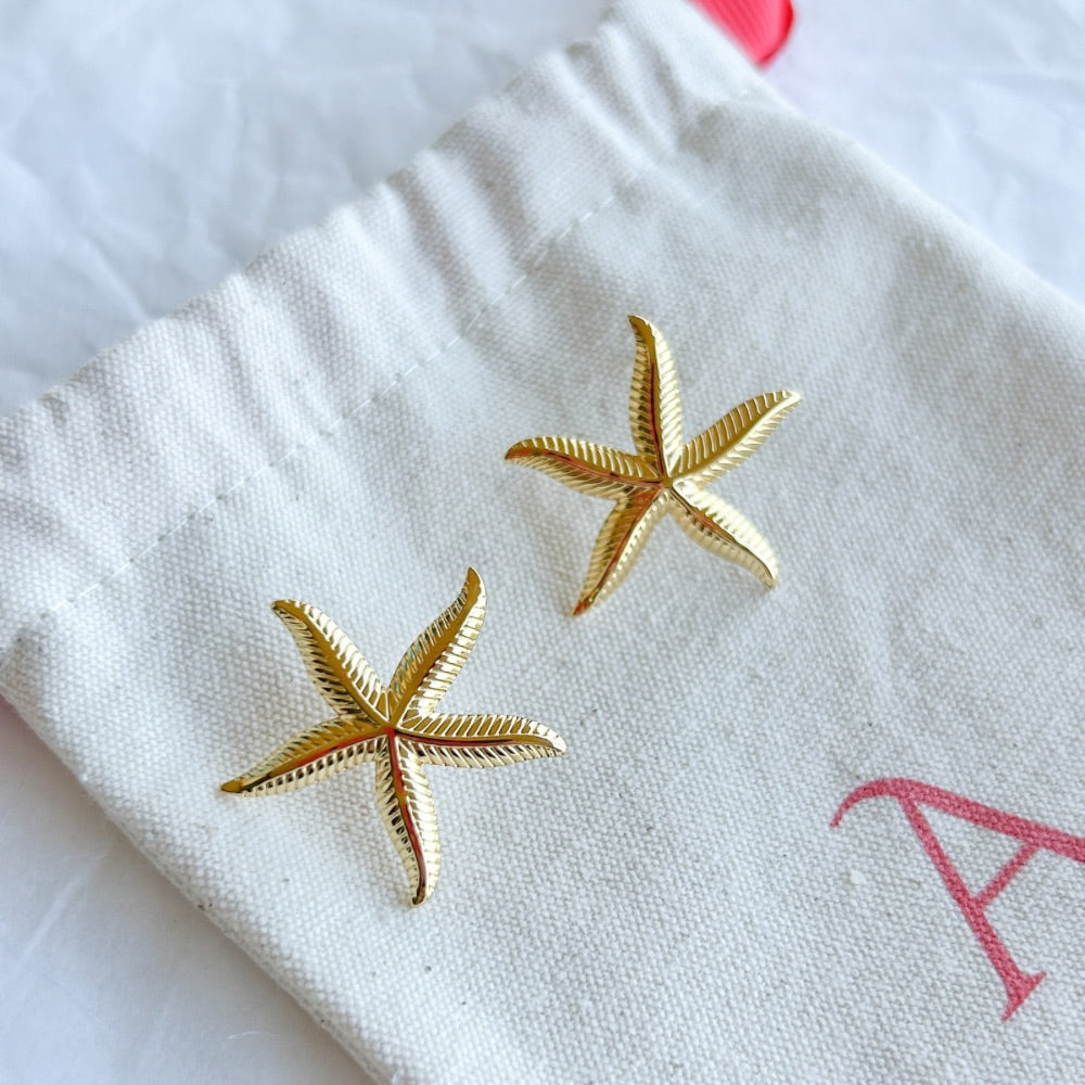 Aros Dorados Aarhus con diseño de estrella de mar y textura detallada, exhibidos sobre una bolsa de tela Altapinta.