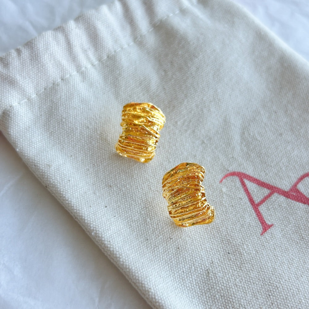 Aros Dorados Bergen con textura entrelazada y acabado en oro brillante, exhibidos sobre una bolsa de tela Altapinta.